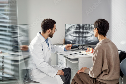 Young male dentist explaining dental x ray results to middle aged Caucasian woman patient in dental office, dentist using pen to indicate details on monitor, patient listening attentively