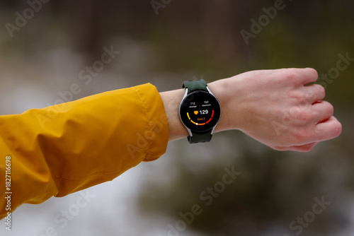 Close-up of an athlete checking heart rate on a smartwatch during a winter workout. The digital screen displays 129bpm and Zone 2 intensity. Person wears a yellow jacket, snowy outdoor environment