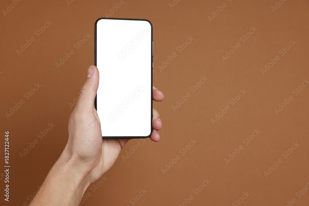 Fototapeta premium Man holding smartphone with blank screen on brown background, closeup. Mockup for design