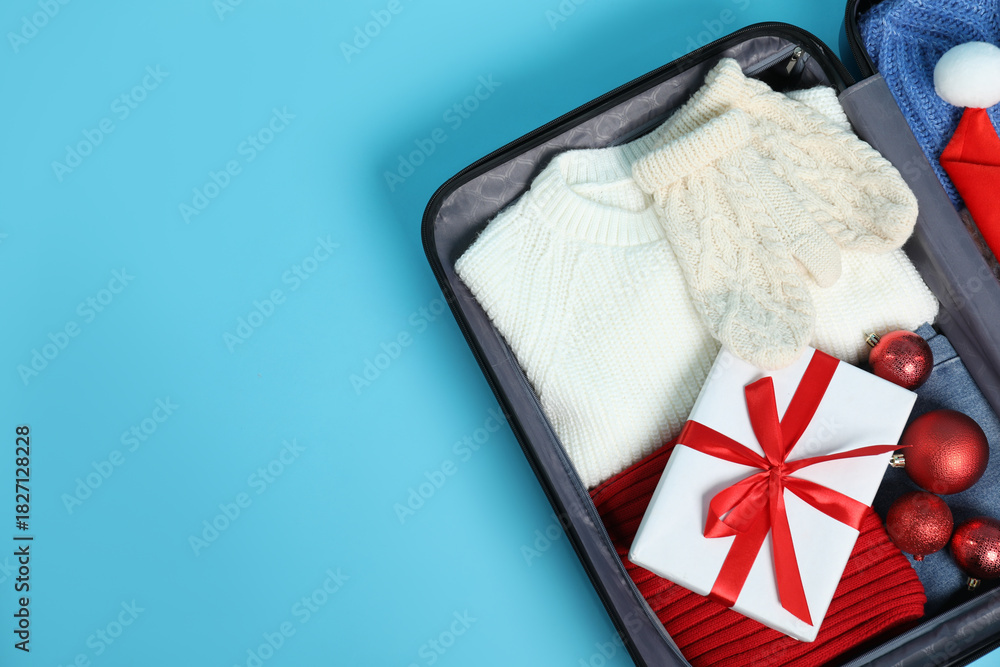 Obraz premium Christmas vacation. Open suitcase with clothes, gift box and baubles on light blue background, top view. Space for text