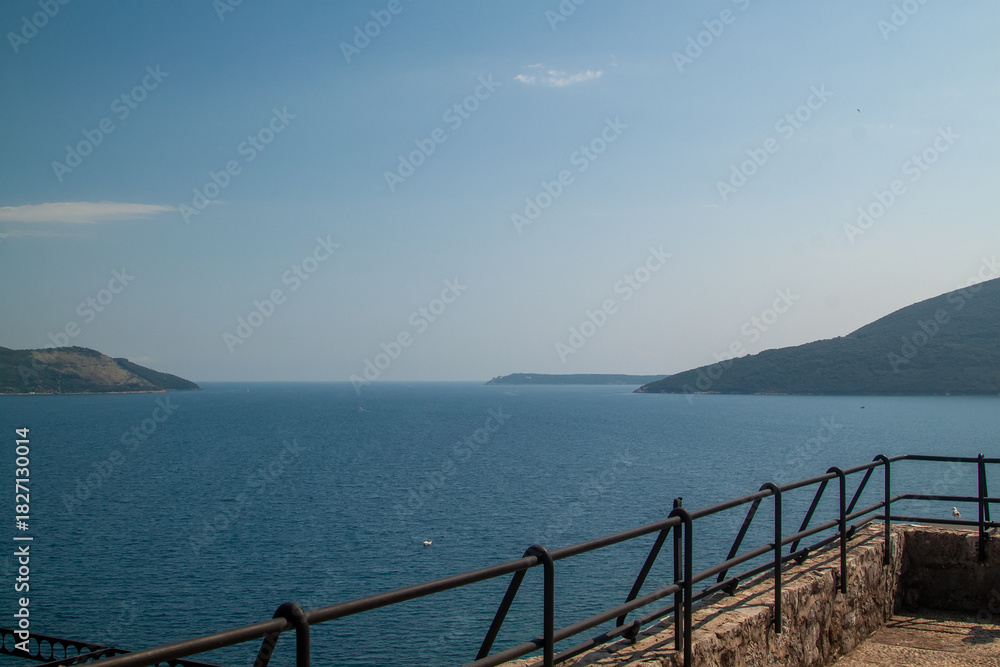 Obraz premium Expansive coastal panorama featuring tranquil blue sea stretching towards distant mountains under clear sky, viewed from stone railing overlook on bright day.