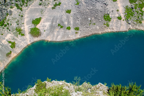Overhead shot of pristine deep blue lake with clear turquoise edges, surrounded by rugged, arid landscape featuring rocky slopes and pockets of green vegetation