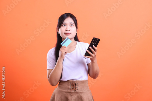 A young Asian woman is holding a credit card and a smartphone while looking up and thinking, about online shopping ideas. isolated on orange background
