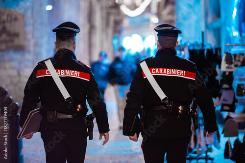 Rear view of two uniformed Italian Carabinieri police officers patrolling a dimly lit, atmospheric, historic street or alleyway in an Italian old town like Bari.