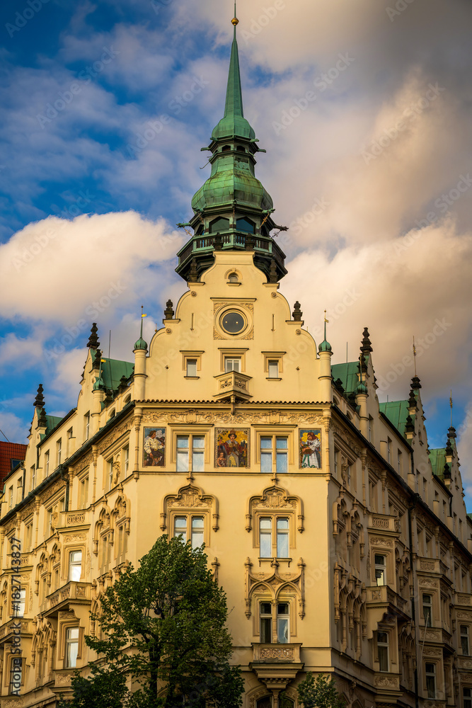Fototapeta premium A grand historic building in Prague featuring a green copper spire, intricate facades, and colorful murals, capturing European architectural charm and cityscape ambiance.
