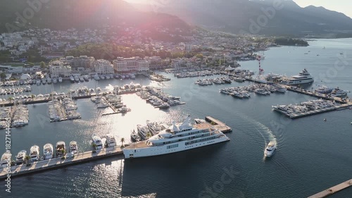Luxury yacht leaving Porto Montenegro marina in Tivat, Montenegro – aerial 4K drone view with superyacht in foreground and luxury waterfront marina with moored boats in the background.