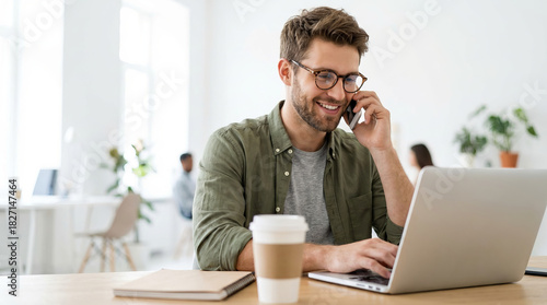 Smiling young businessman talking on the phone while working on his laptop in a bright, modern corporate office space.