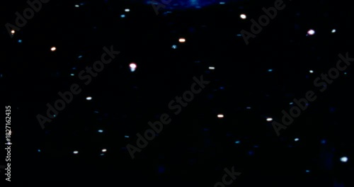 Red, blue, and silver particles drift through water in a dark space, reflecting cool and warm light—cosmic, dreamy, glittering star-like movement.