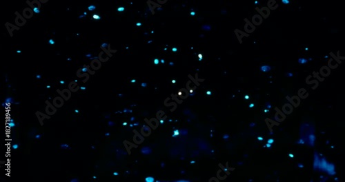 Particles float in water over a dark background, reflecting top light in green and blue hues, creating a glittering, halo-like, cosmic universe effect.