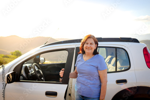 Smiling mature Brazilian woman standing by her car on a sunny trip, celebrating independence, confidence, and the freedom to travel after 60.