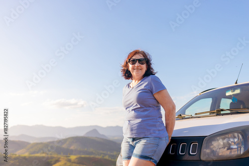 Brazilian mature woman enjoying a peaceful road trip, smiling beside her car with a scenic mountain view. Inspiring independence, freedom, and confidence for women over 60