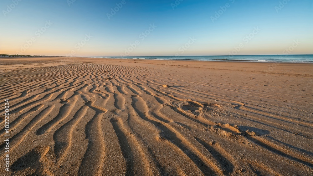 Naklejka premium A sandy beach with rippled patterns, clear blue sky, and calm ocean waves. Nature and landscape, tranquility and serenity, outdoor scene.