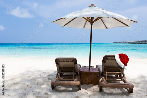 Fototapeta Naklejka Na Ścianę i Meble -  Santa Claus hat on tan chairs under white parasol on turquoise sea beach, perfect vacation on Maldives island. Christmas and New Year celebration