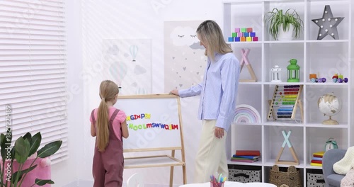 Speech therapist teaching girl alphabet near whiteboard with letters indoors