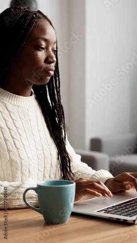 A woman with long braids uses a laptop while holding a mug in a cozy indoor setting