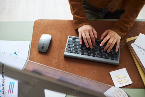 Young Asian woman working at office desk typing on wireless keyboard with hands visible, surrounded by business documents and notes, engaging in professional business tasks