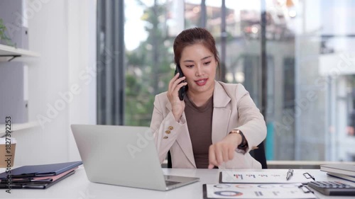 Asian business professional woman multitasking phone call laptop office desk analyzing data corporate communication