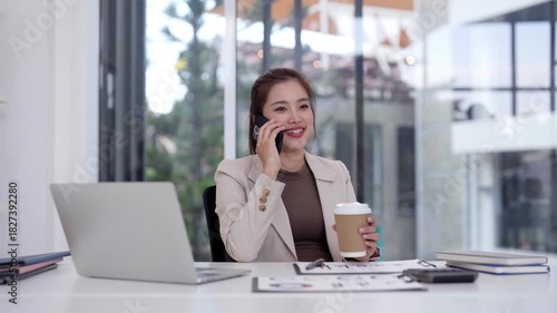 Asian Business Woman On Phone Call Managing Work Tasks With Laptop In Modern Office Environment