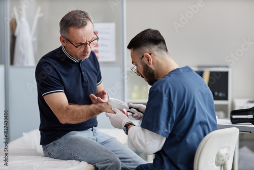 Middle aged Caucasian man sitting on examination table receiving skin check from young adult male dermatologist using magnifying glass in medical office