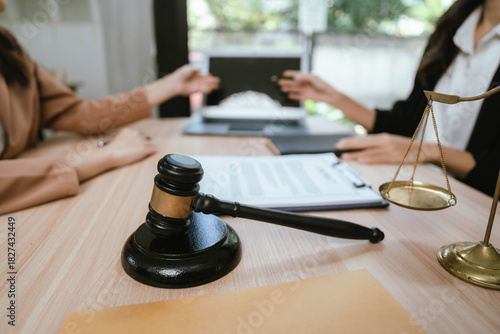 Two professional women discussing legal documents at an office desk, reviewing contracts with a laptop and gavel nearby.
