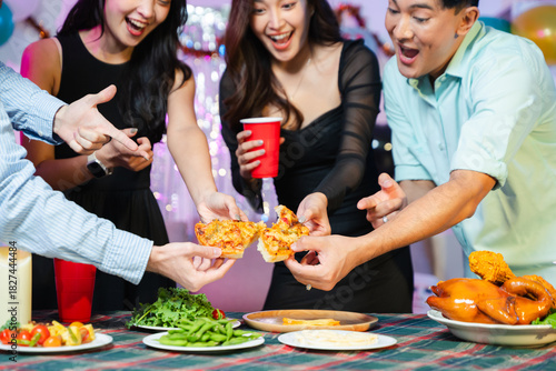 Hands of cheerful friends holding and sharing pizza slices together at a festive indoor party table with snacks, red cups, and celebration vibe