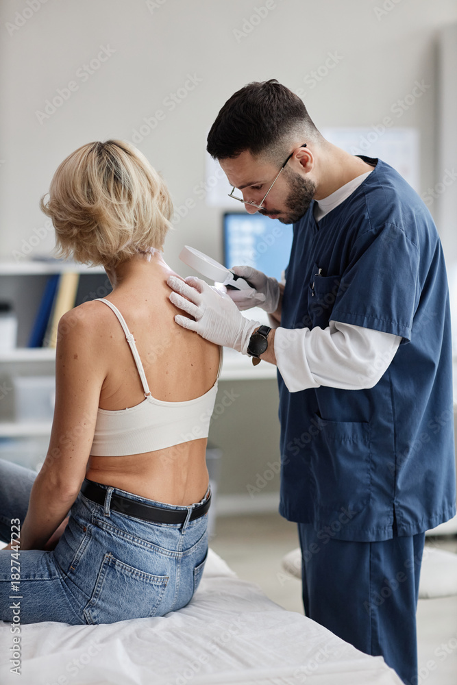 Fototapeta premium Caucasian woman sitting on examination table receiving skin check from Middle Eastern young adult male dermatologist wearing medical gloves and using magnifying glass
