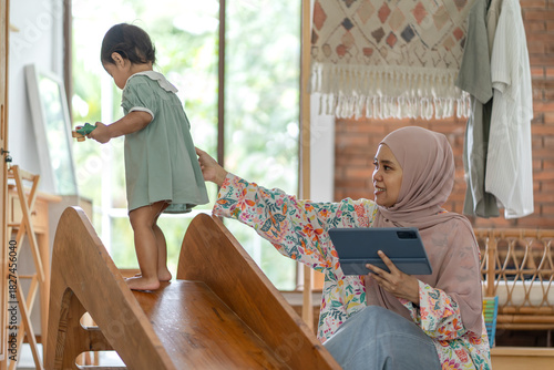 Indonesian Muslim Woman Remote Working on Tablet with Daughter Playing Indoors