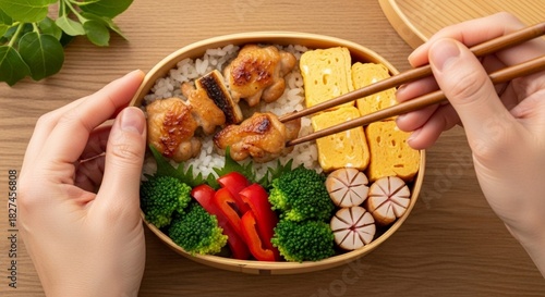 Overhead shot of a bento box filled with rice, chicken, vegetables, and egg, held by hands using chopsticks.