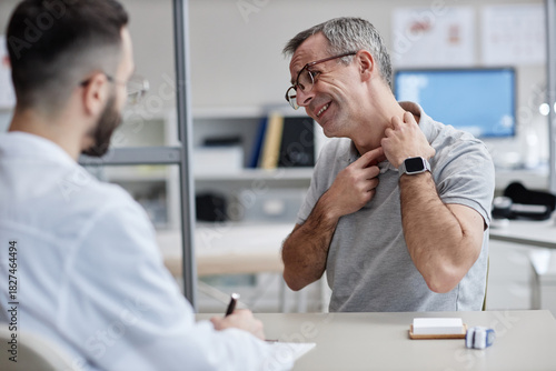 Middle aged Caucasian man describing skin irritation on neck while consulting male doctor in medical office, patient showing symptoms during dermatology appointment, healthcare professional listening