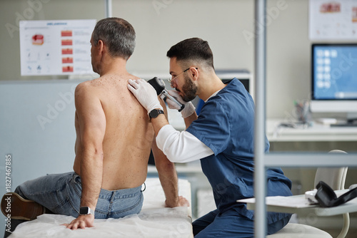 Middle aged Caucasian man sitting on examination table being examined by young adult Caucasian male dermatologist using dermatoscope during skin check in medical office