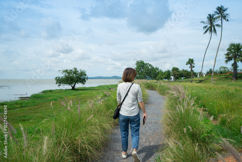 A woman to walk alone and see view of sea with along gravel walkway and flower grass at the park.