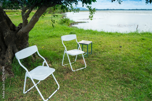 Chairs and table on the grass lawn near the lake with cloud and blue sky background . Our door living to meeting.