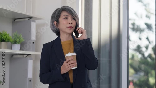 Asian older businesswoman checks smartwatch works laptop manages time schedules tasks boosts productivity modern office professional workspace