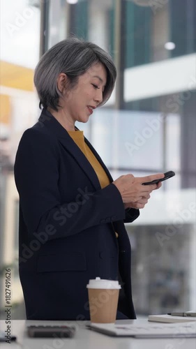 Asian senior businesswoman talking mobile phone using laptop managing business tasks in professional modern office communicating