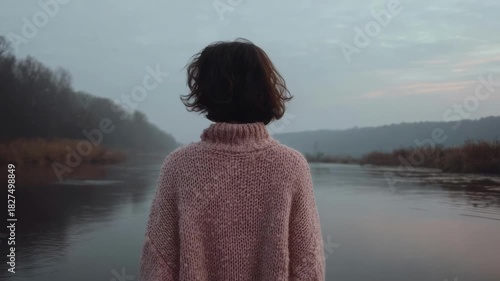 Back view of a person in a pink chunky knit sweater standing by a calm, misty river at dusk.