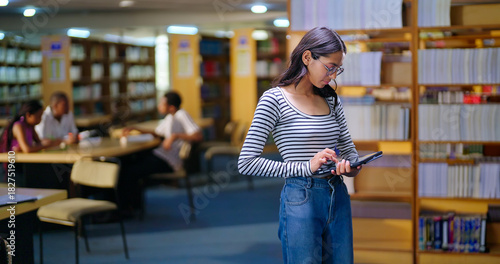 Indian young adult gen z busy woman lady hold tab look screen do online work e learning skill check exam notes. Happy teen girl wear glass candid casual pose enjoy new day time indoor library