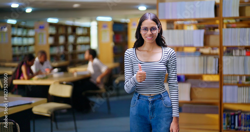Indian young adult gen z happy student wear glasses indoor library look camera show thumbs up hand sign ready for exam Asian teen age girl staring cam do casual pose enjoy day time inside college