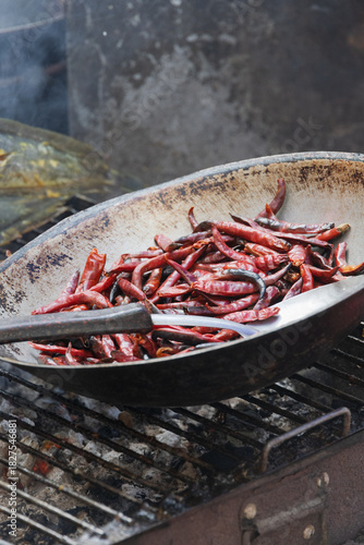 Dried red chili peppers fry in a rustic wok over a smoking dark metal grill in Bangkok.