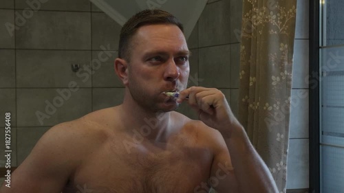 A man's morning, getting ready for work. Morning hygiene, brushing his teeth in front of the mirror, the man looks sleepy.