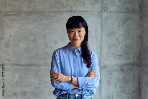 Asian Woman in Blue Shirt Stands Confidently With Crossed Arms in Modern Workspace