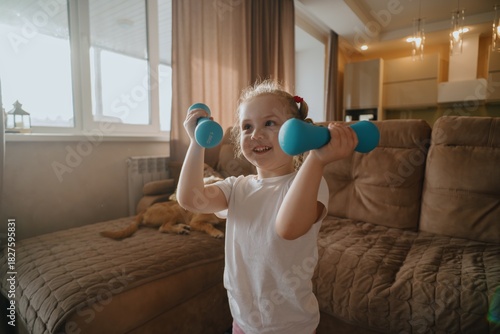 A joyful 4-year-old girl does morning exercises in a bright living room. Authentic moment of child fitness, energy and healthy family routine