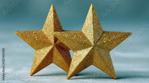 Two glittery golden stars stand side-by-side against a muted blue-gray background. The stars are detailed and textured, creating a sparkling effect.