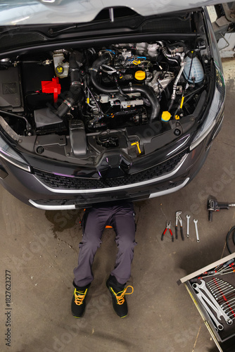 Top view of a mechanic working under a lifted car. Open hood and tools on the floor for vehicle maintenance and engine repair in the garage.