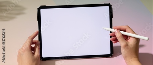 Top view of hands holding tablet with blank screen and white stylus on pink surface.