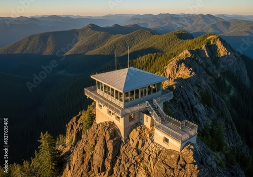 A modern, white observation deck with a flat roof and large windows sits atop a rocky cliff, overlooking a vast, forested mountain range