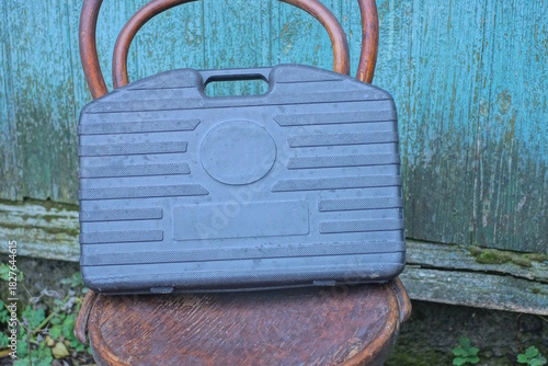 One large black closed plastic toolbox stands on a brown chair against a green wall on a summer street