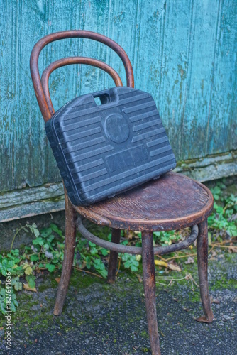 One large black closed plastic toolbox stands on a brown chair against a green wall on a summer street