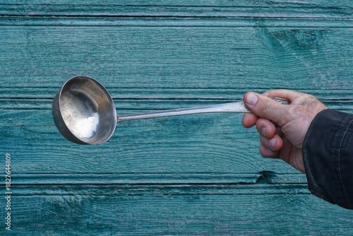 hand holding one big gray old metal ladle spoon against green wall background outdoors