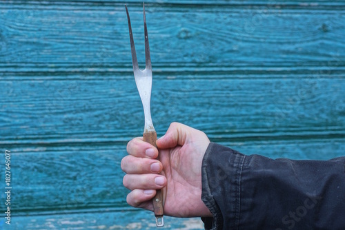 A man's hand in black clothes holds one large metal fork with a wooden handle against a green wall on the street