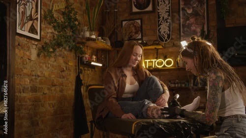 Handheld shot of professional young Caucasian female tattooist with dreadlock hairstyle using tattoo machine while working with female customer in cozy modern studio
